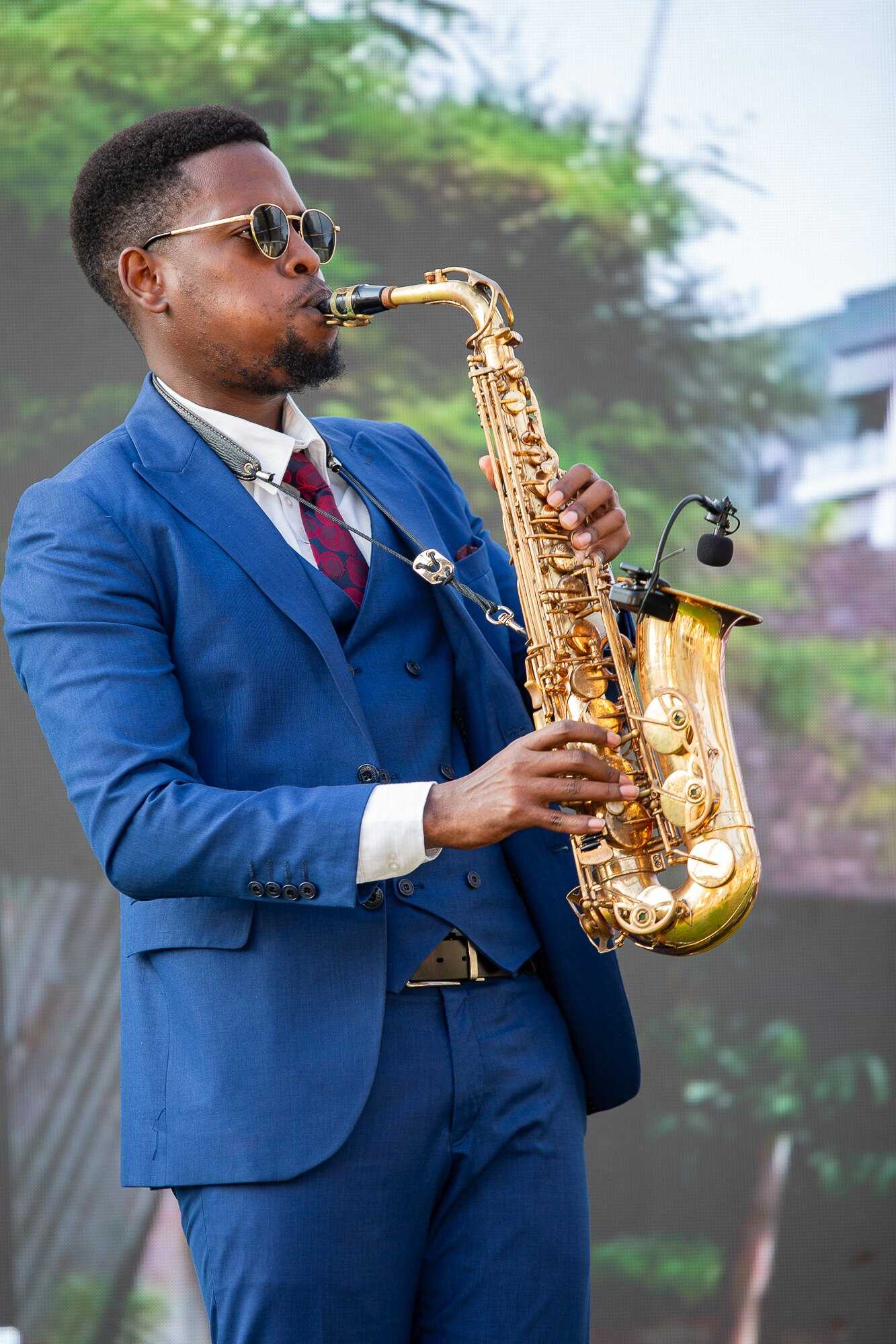 Close portrait of Ian in a blue three-piece suit and round sunglasses, playing a gold alto saxophone with a wireless mic