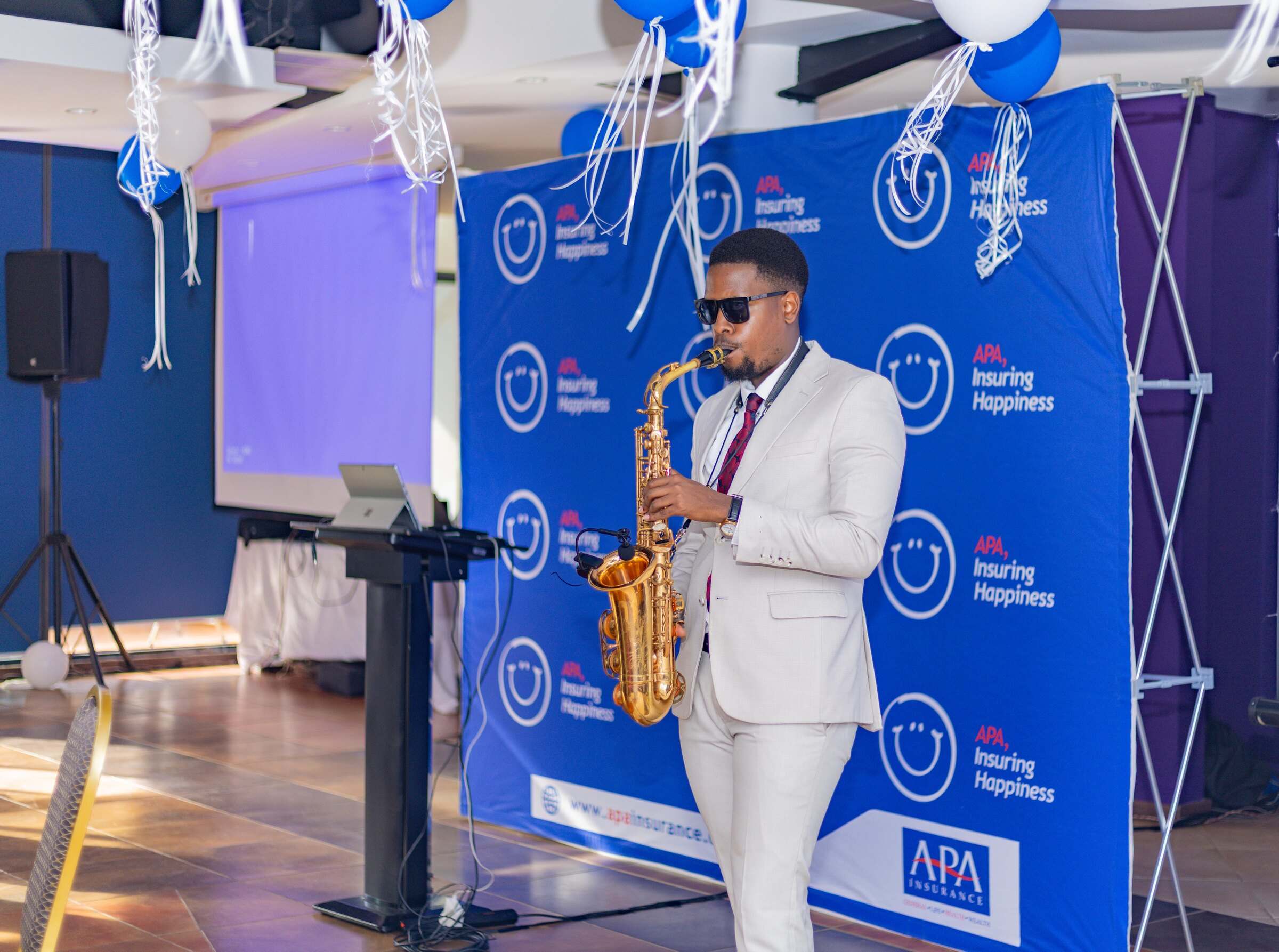 Ian in a cream suit playing saxophone in front of a branded backdrop at a corporate event, balloons overhead