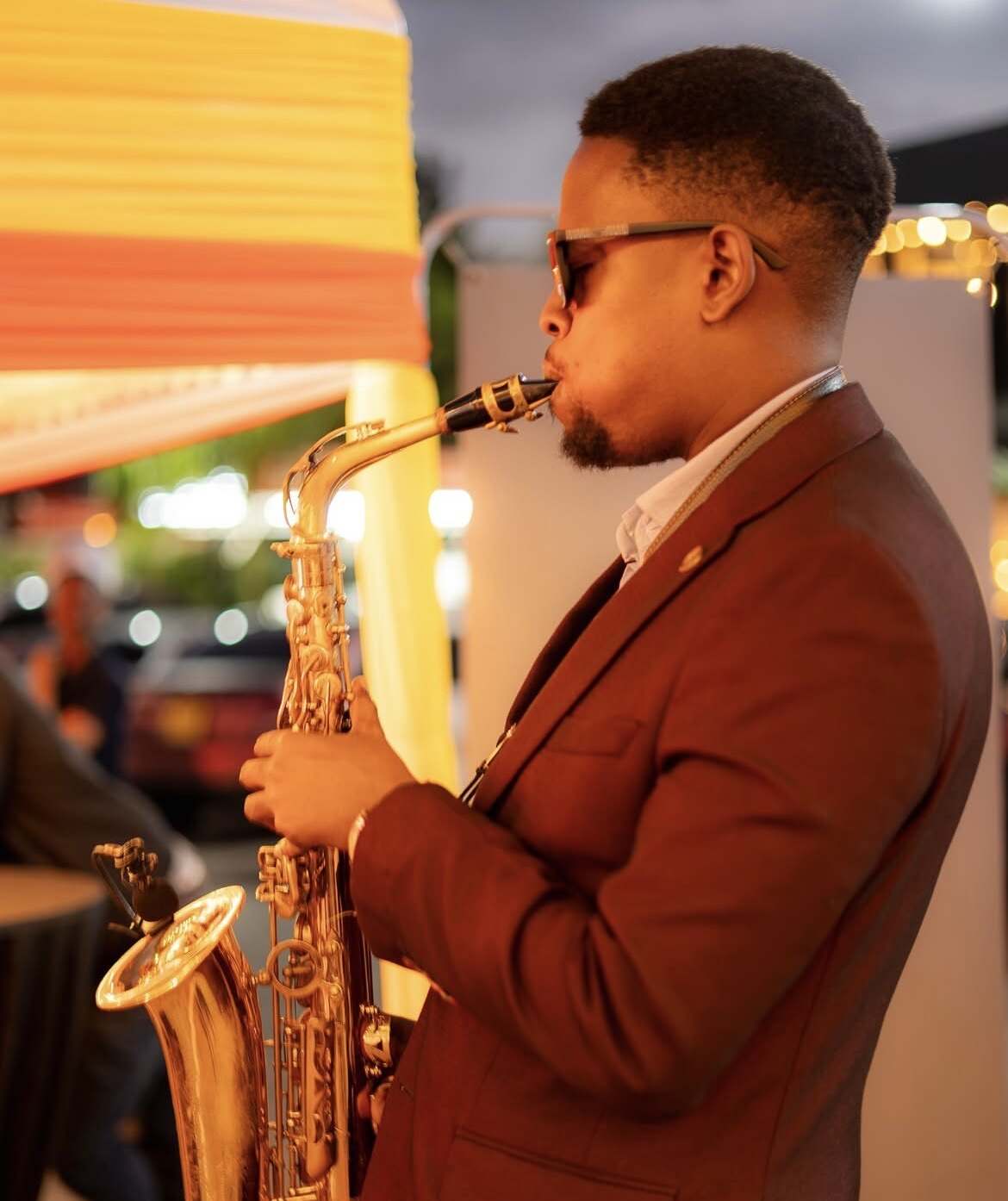 Side profile of Ian in a rust-red blazer playing saxophone at an evening event strung with fairy lights