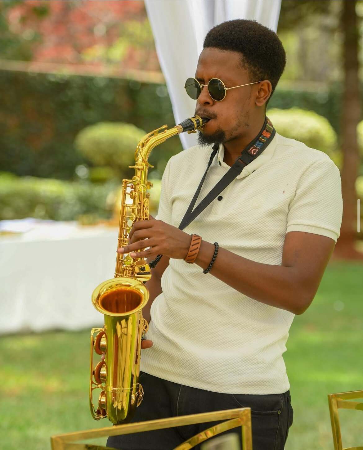 Ian in a white knit polo playing a gold saxophone at a sunny garden party