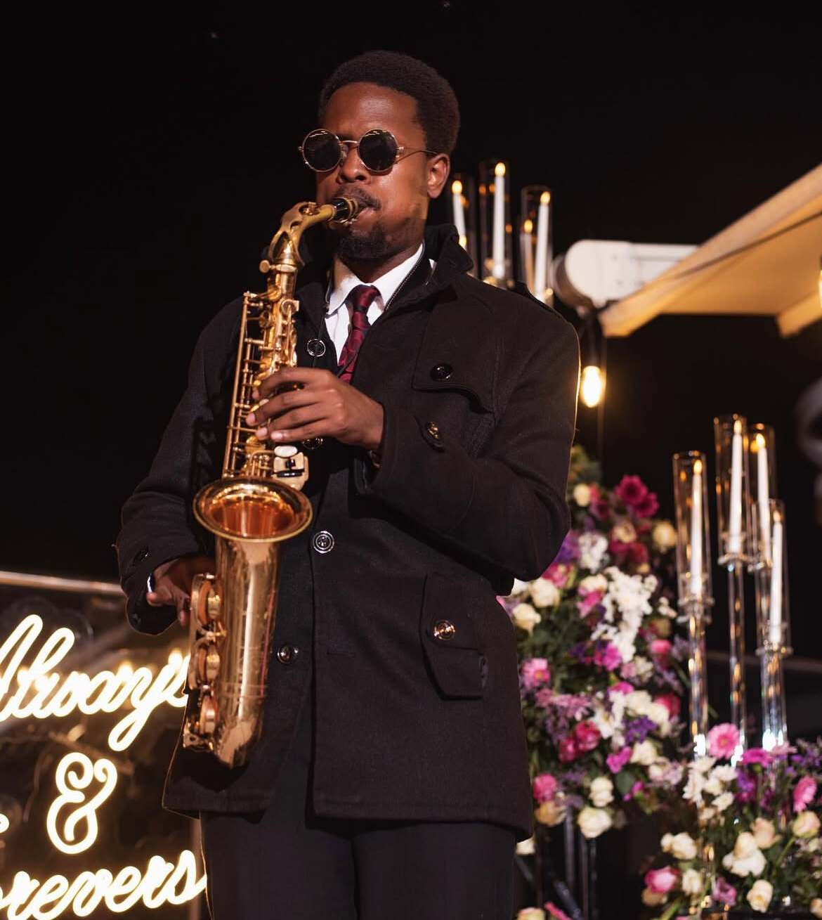Ian in a dark coat playing saxophone at a night-time proposal set with tall candles, flowers and a neon sign