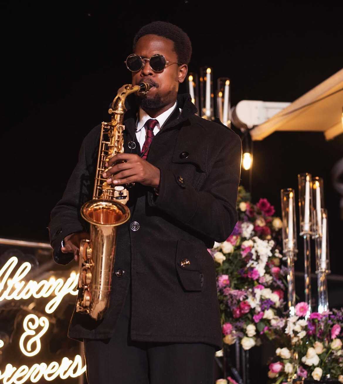 Ian in a dark coat playing saxophone at a night-time proposal set with tall candles, flowers and a neon sign