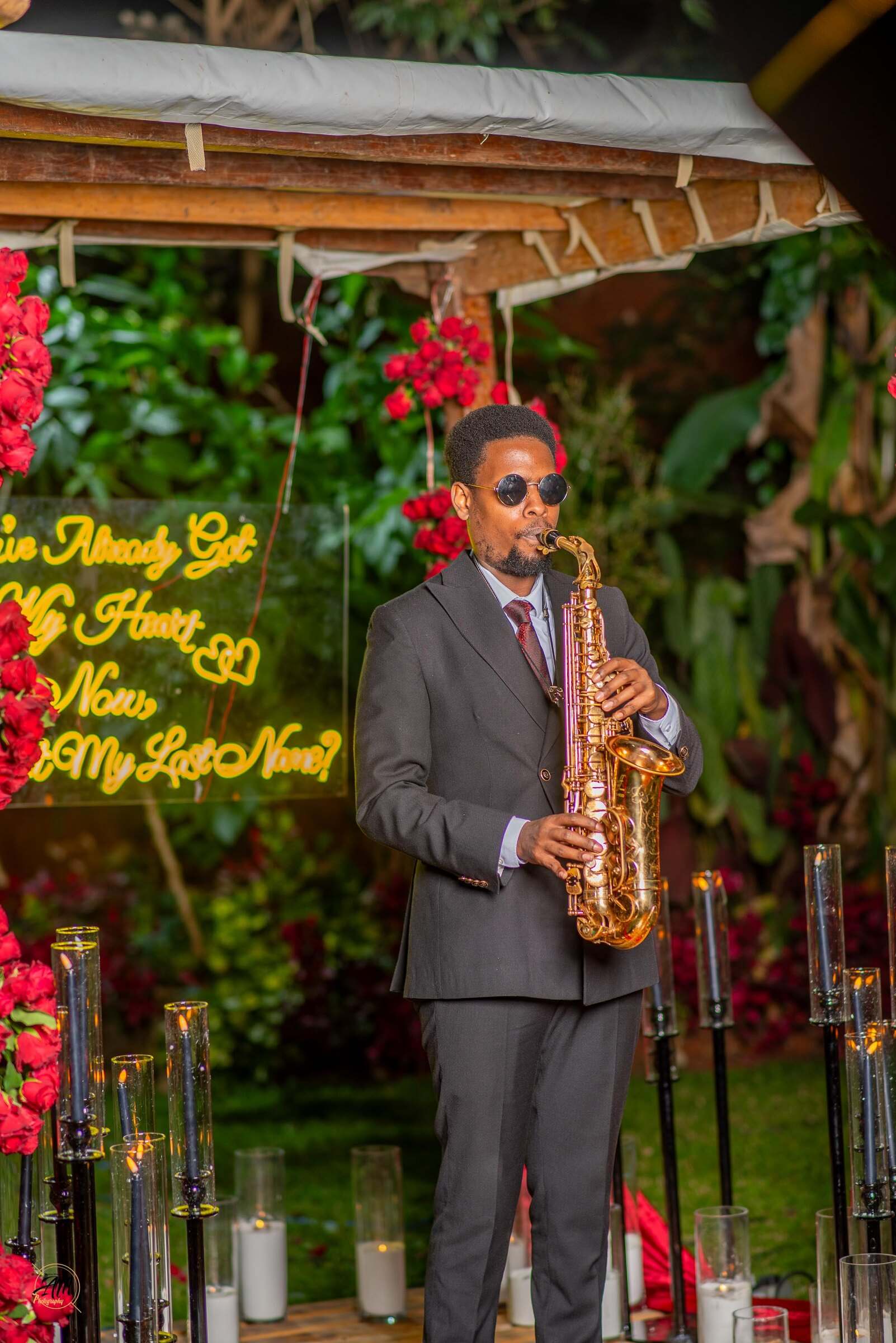 Ian in a grey suit playing saxophone at a garden proposal set framed by red roses, candles and a neon sign