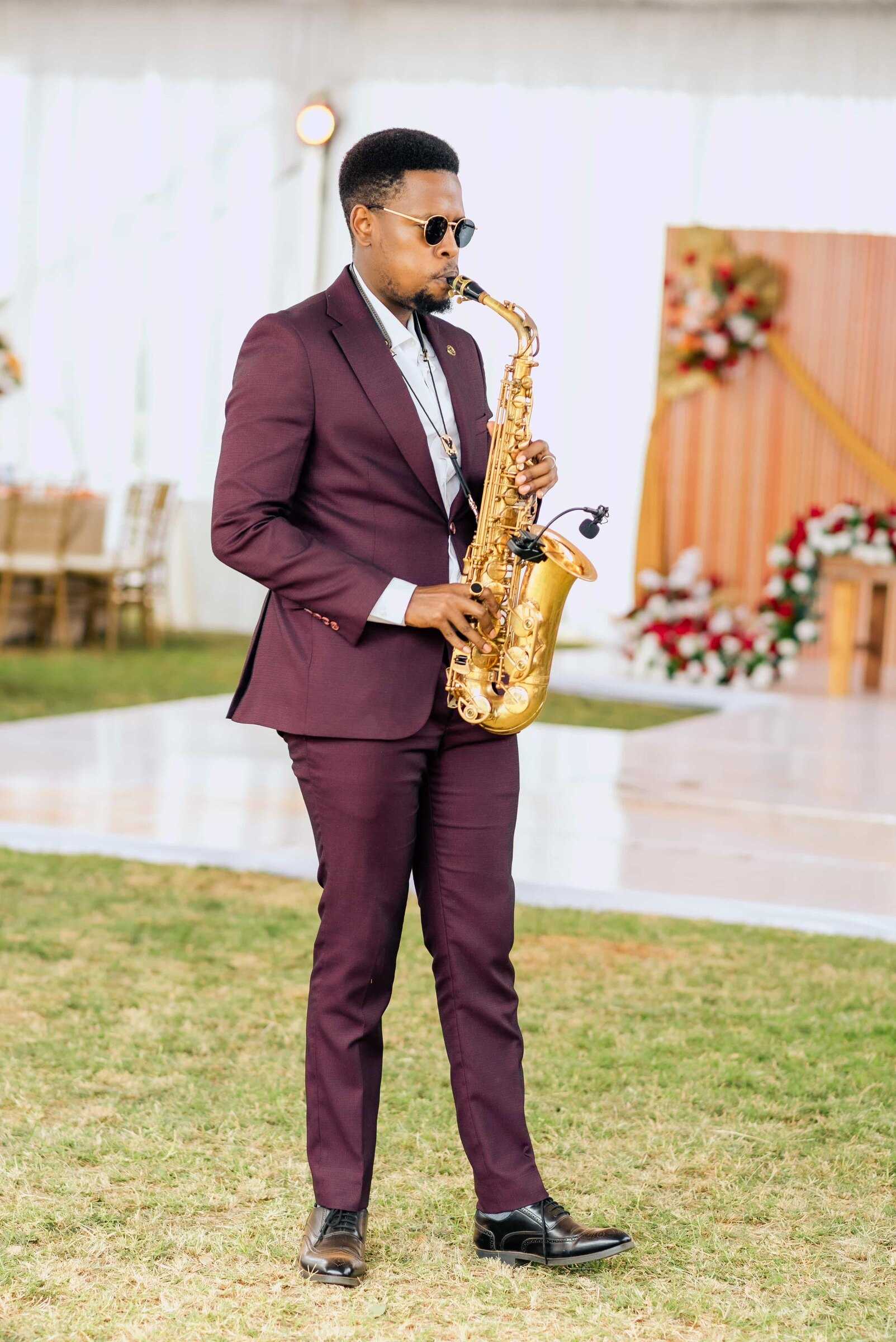 Full-length view of Ian in a burgundy suit playing saxophone on the lawn beside a wedding dance floor