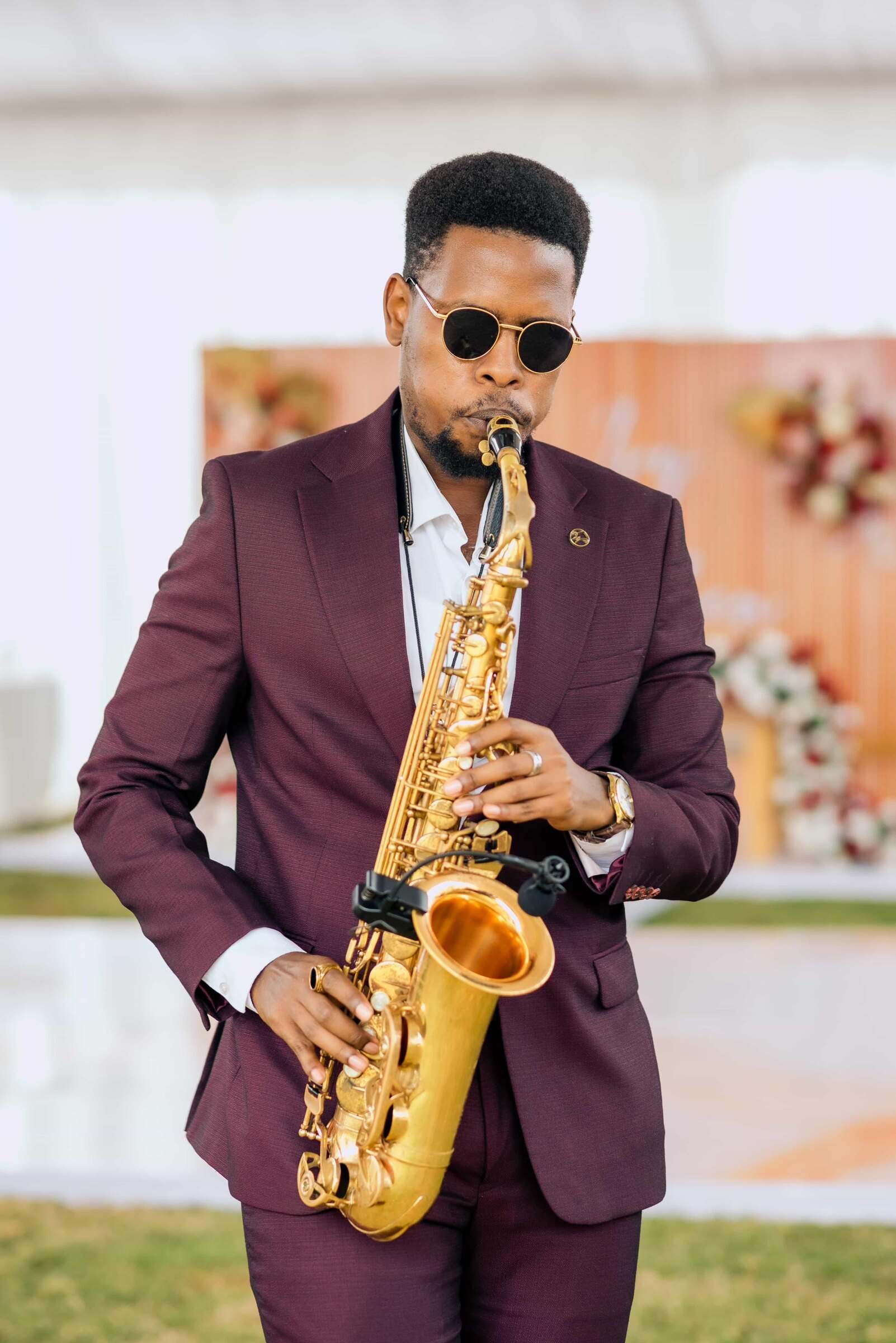 Ian in a burgundy suit and round sunglasses playing a gold saxophone with a clip-on wireless mic at an outdoor wedding