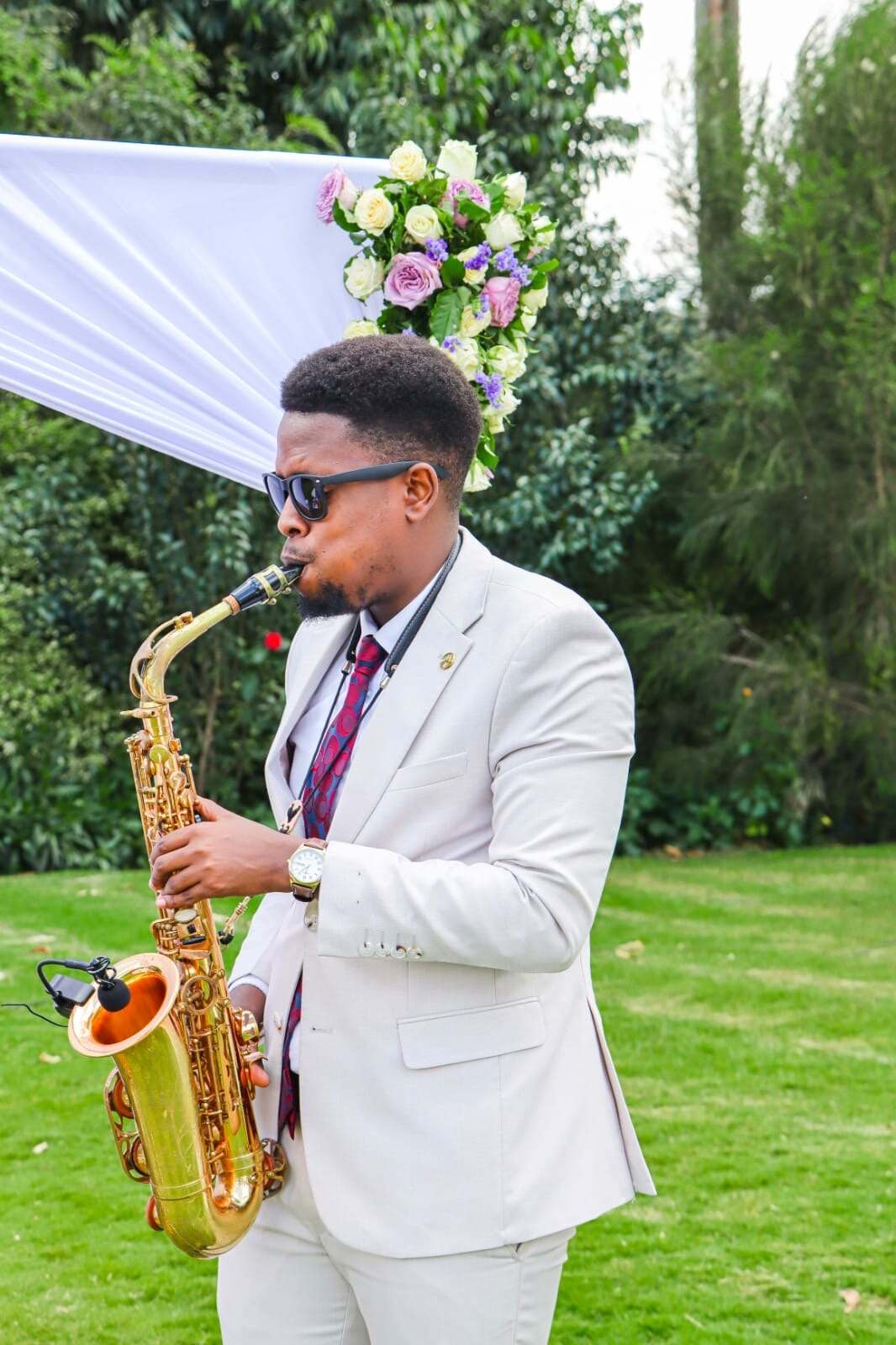 Ian in a cream suit playing saxophone beside a floral arch on the lawn at an outdoor wedding ceremony